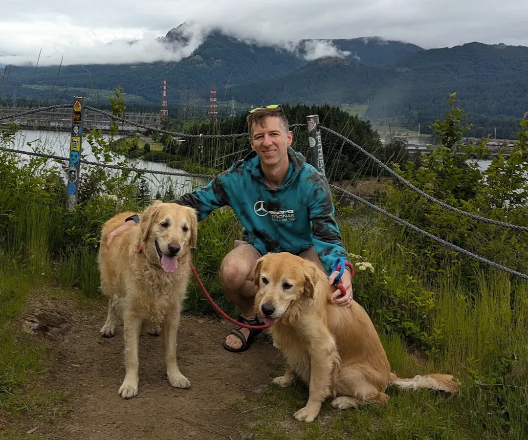 Brandon Cantrell crouching between his two golden retrievers on a trail above the Columbia River Gorge